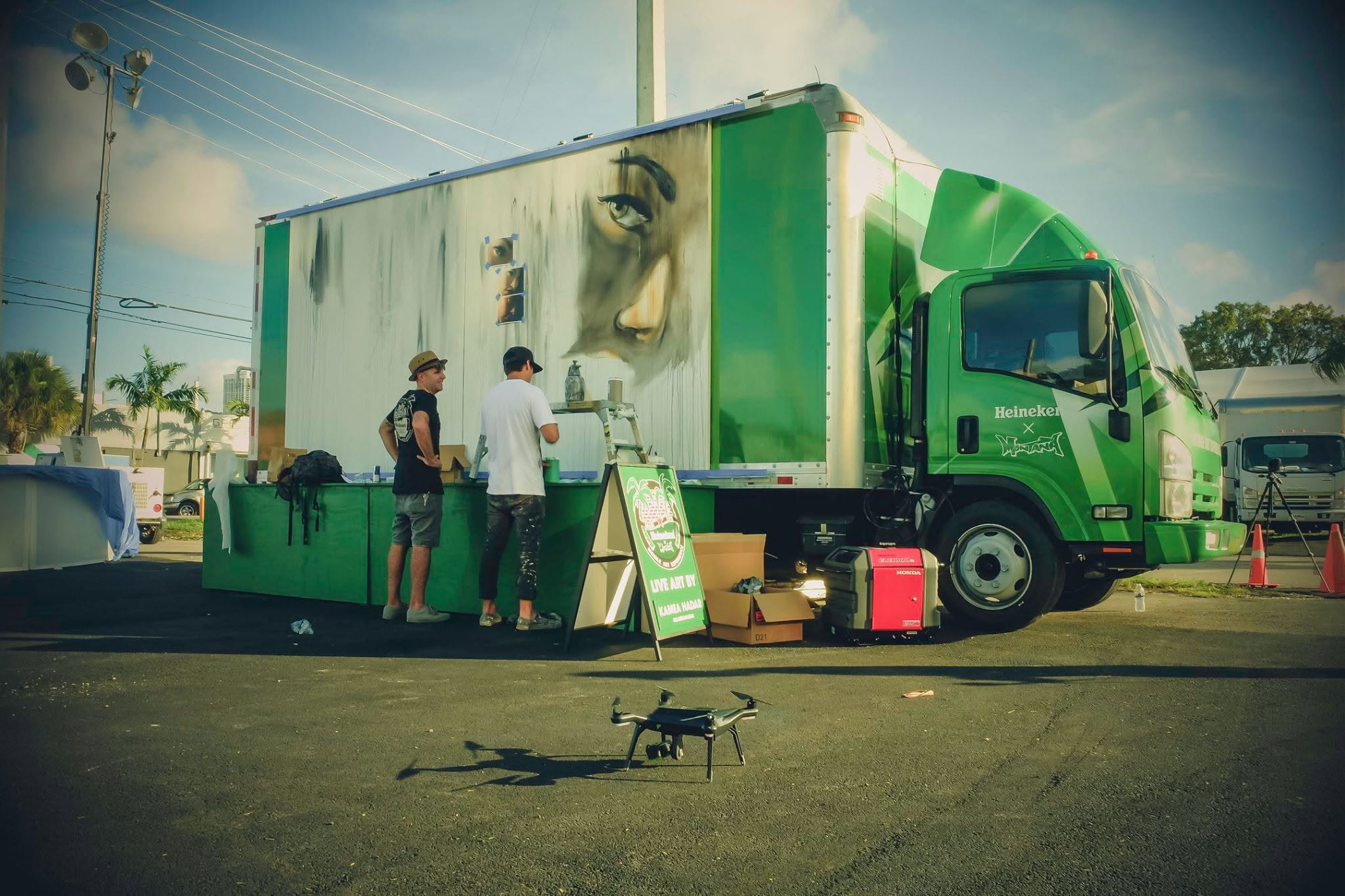 A green Heineken × Montana truck with a half-painted portrait mural on its side, two people working beside it, and a drone resting on the pavement in front.