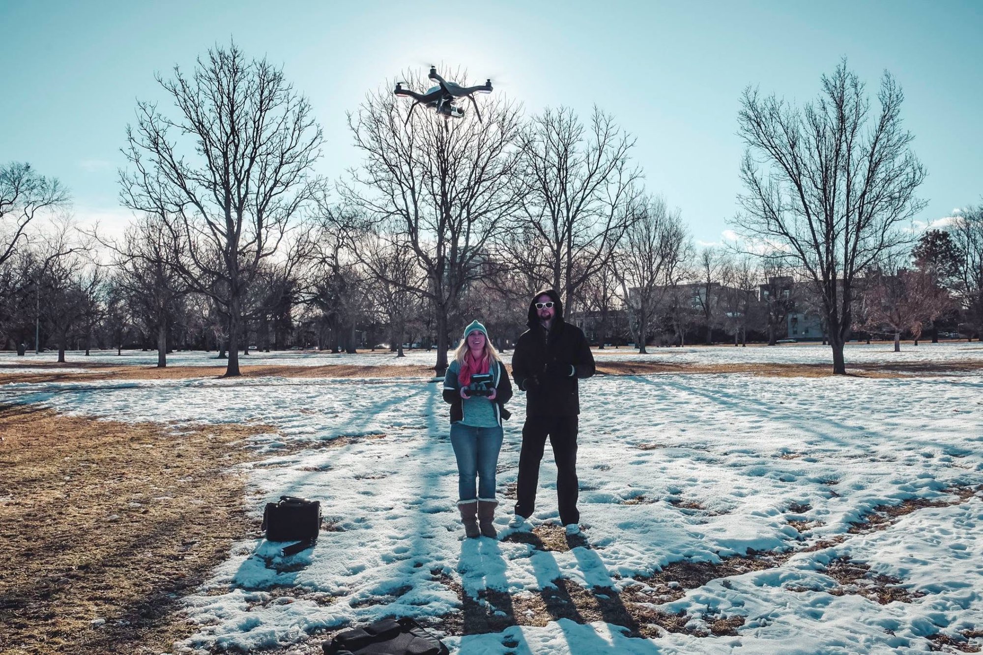 Aviator Marisa Jarae and another pilot stand in a snowy park, with a drone flying overhead.