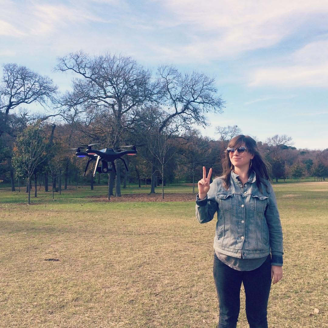 Kate in a denim jacket and heart-shaped sunglasses makes a peace sign in a park as a drone hovers beside her.