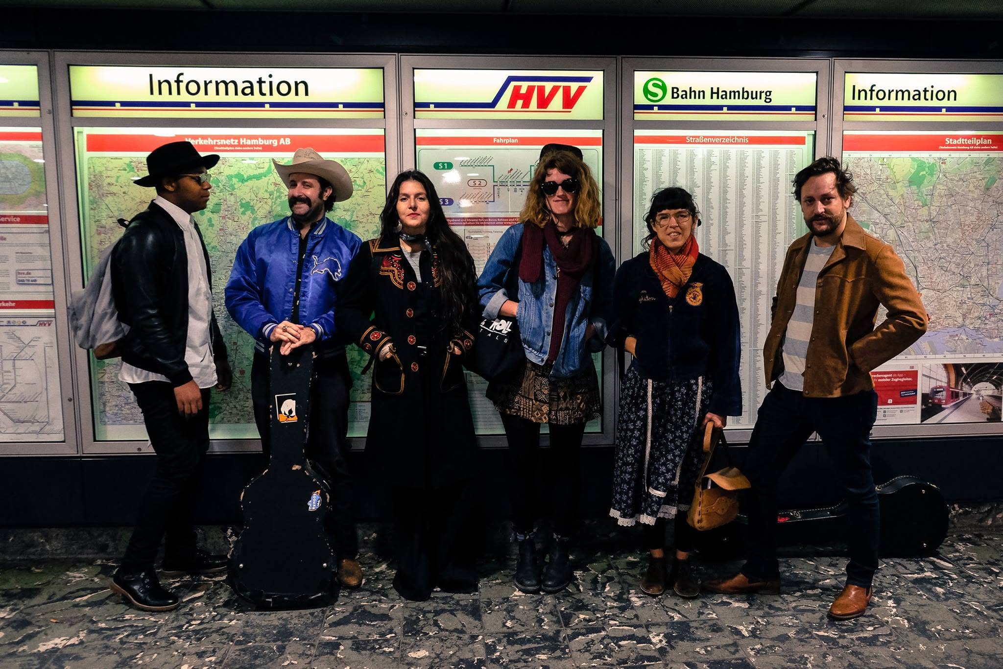 Six musicians with instrument cases standing in front of the S-Bahn Hamburg information maps in a train station.
